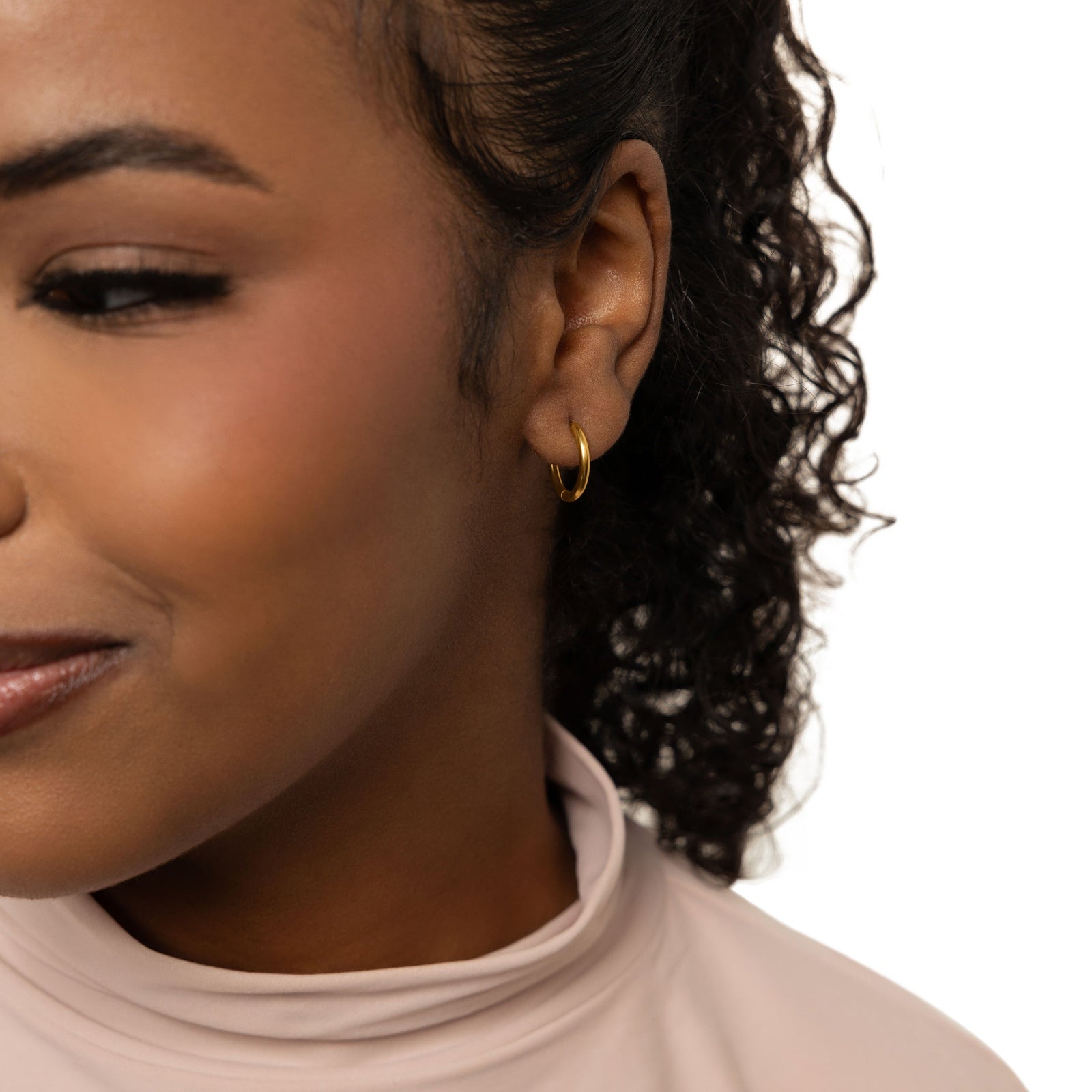 The gold Huggie Essential Hoops | 12mm as worn by the model on their left ear.