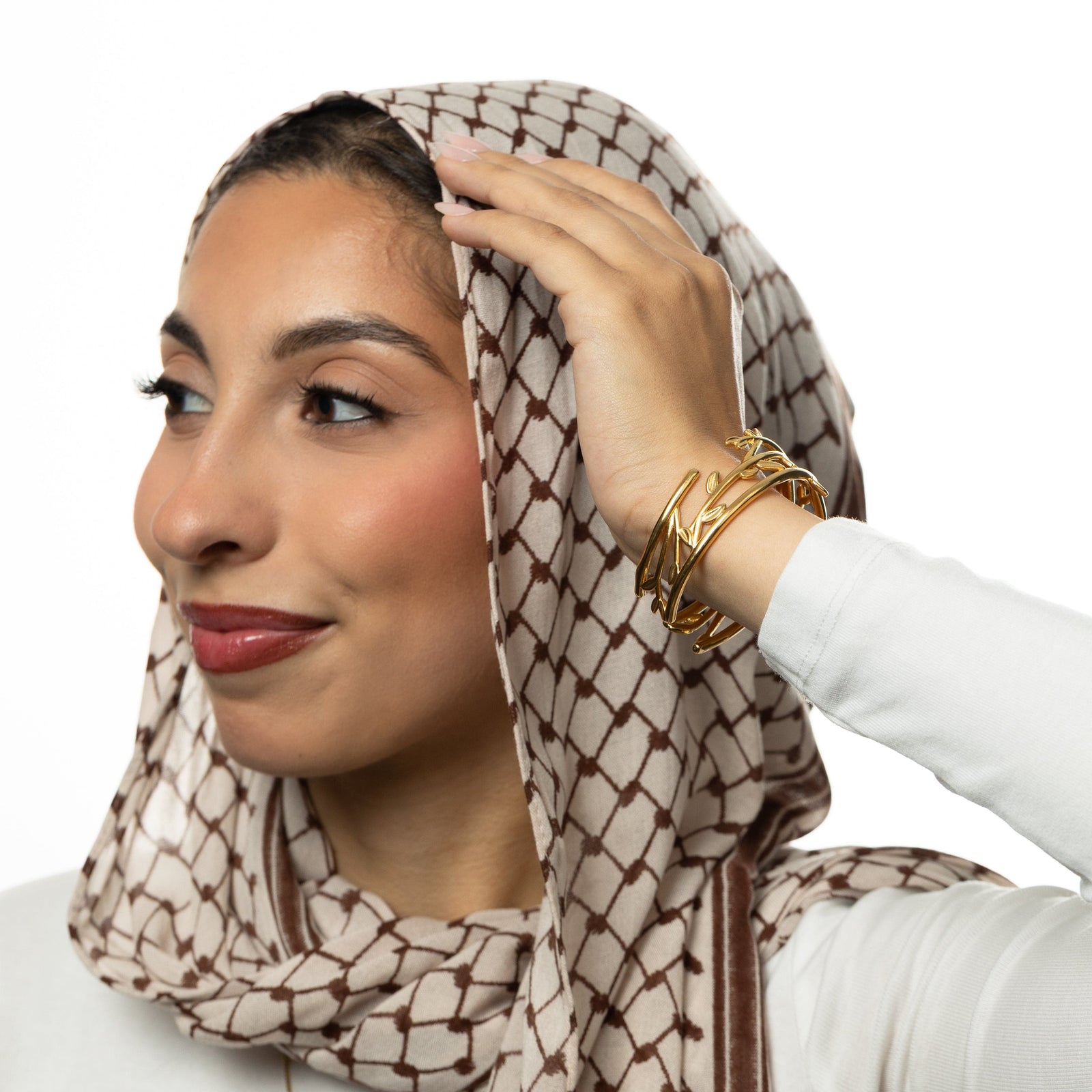Woman with hand on her keffiyeh patterned headscarf, wearing gold Imperfect Olive branch cuff on wrist.