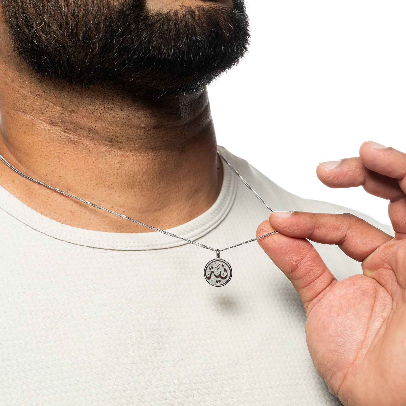 Man wearing a silver intention token necklace, gently pulling the necklace, dressed in a white shirt.