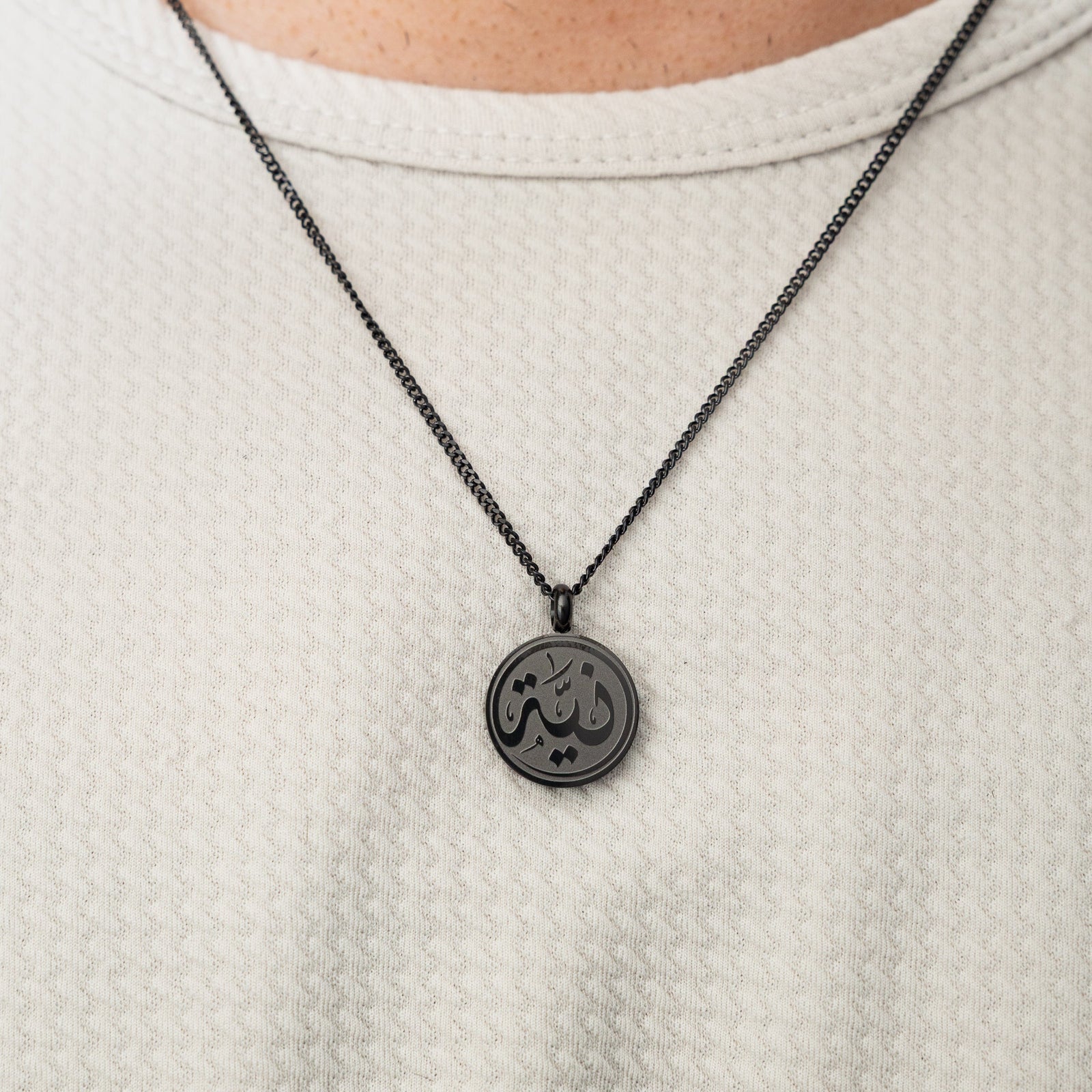 Black intention token necklace worn over a white textured shirt.