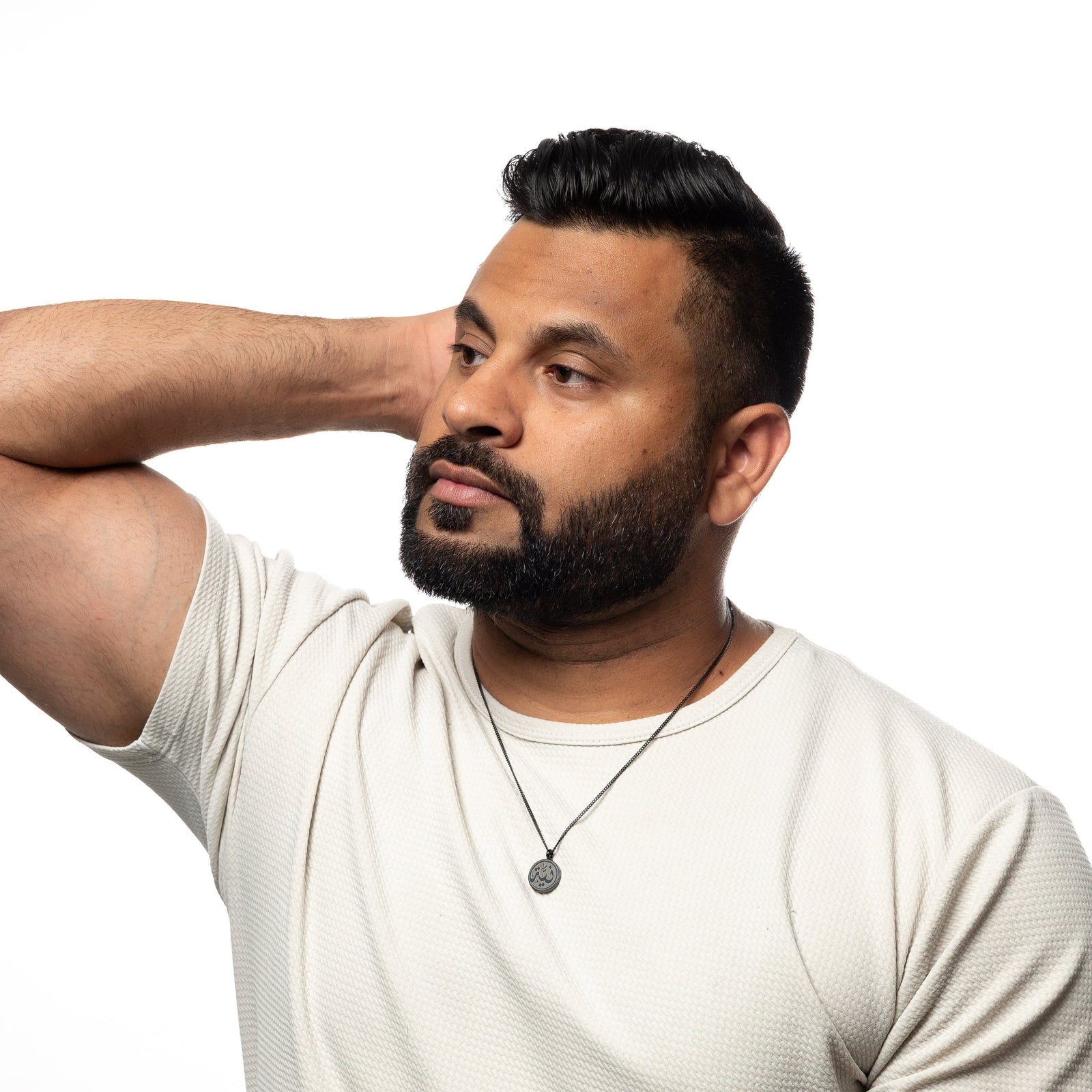 Man wearing a black intention token necklace, worn over off-white textured shirt, hand resting behind his head.