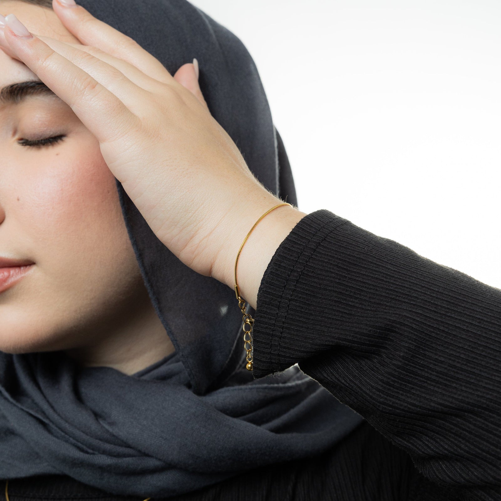 Photo of the model touching their dark gray hijab with their left hand. The model is wearing the gold Micro Harmony Bracelet on their left wrist.