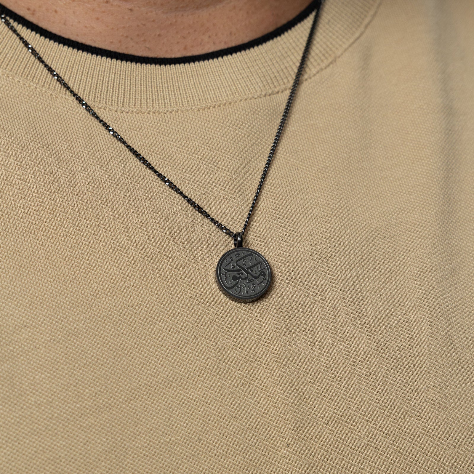 Black 'Maktub' token necklace worn over a beige knit shirt.