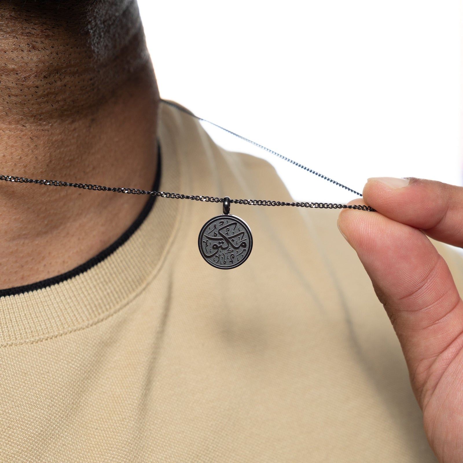 Man holding up a black necklace with a circular pendant engraved with 'Maktub' calligraphy, worn over a beige shirt.