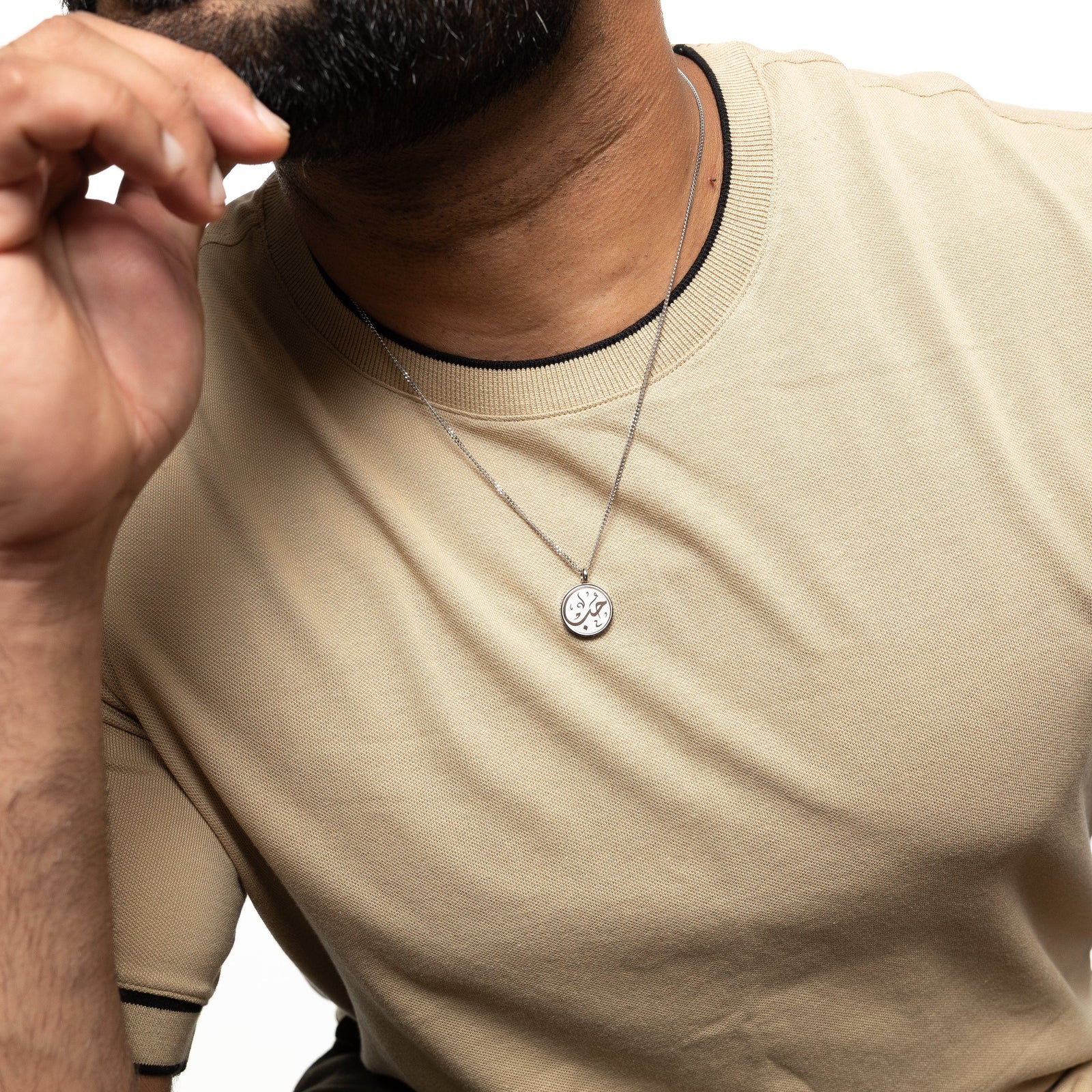 Man wearing a silver 'Maktub' token necklace, resting his hand near his chin.