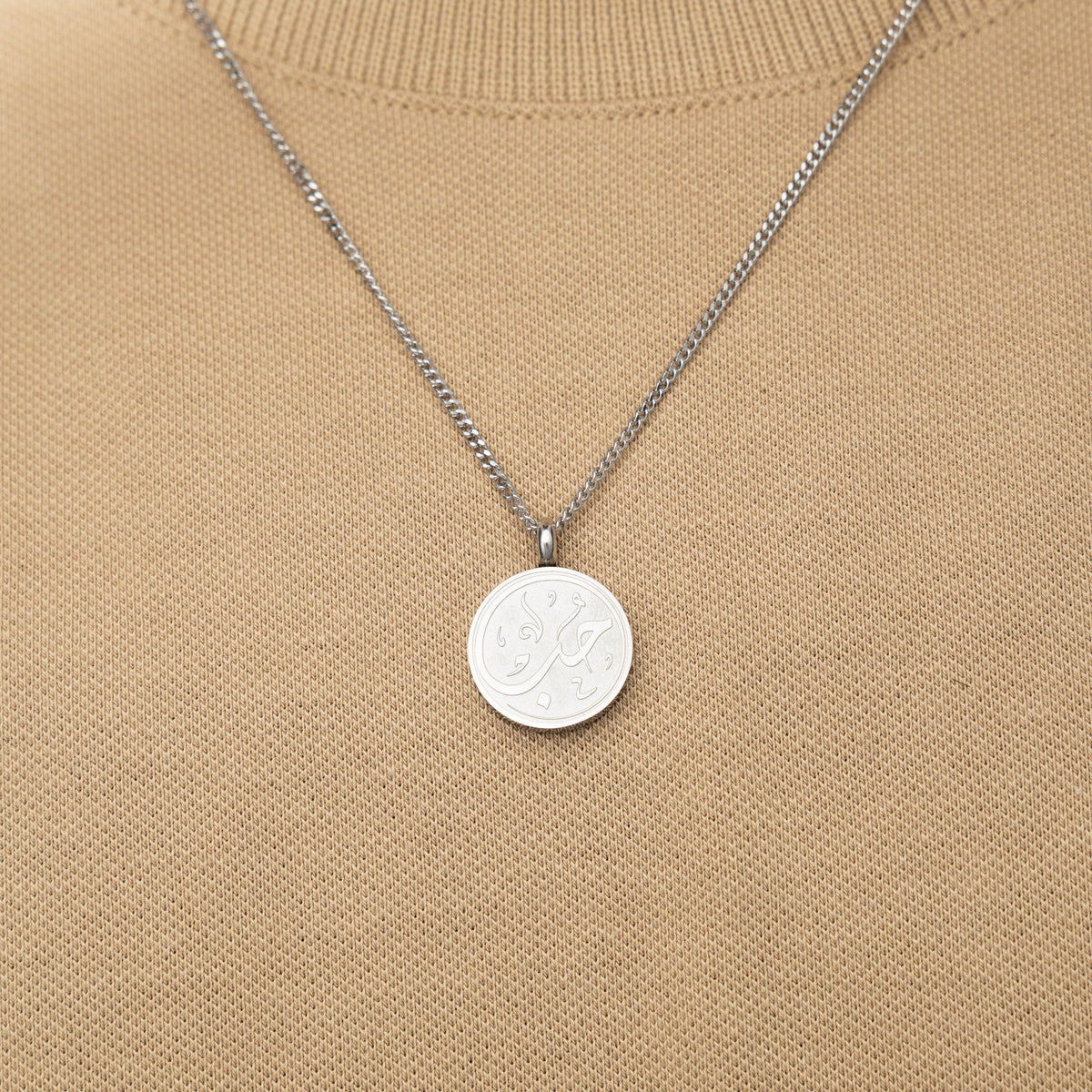 Silver necklace with a circular 'Maktub' calligraphy engraved-pendant worn over a beige knit top.