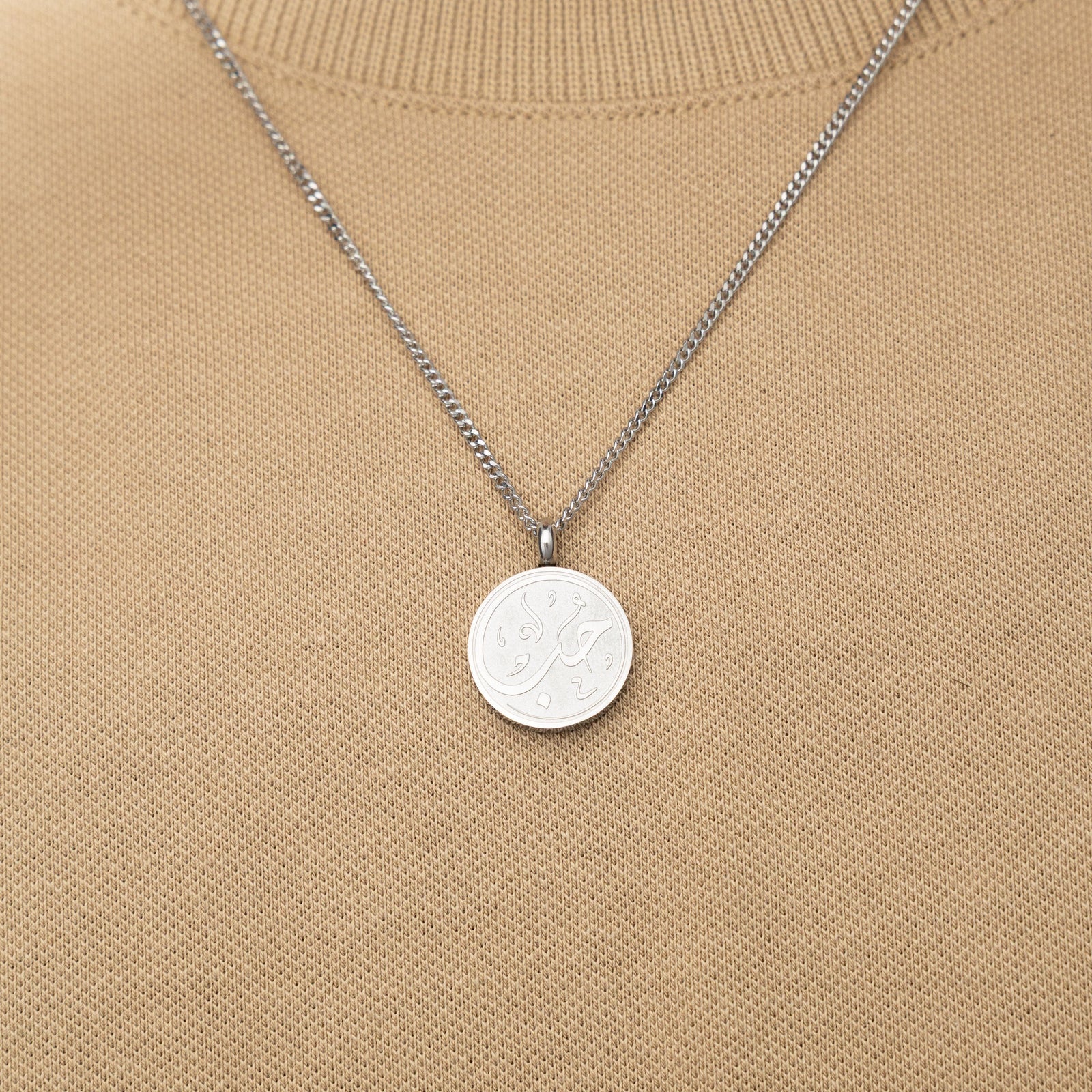 Silver necklace with a circular 'Maktub' calligraphy engraved-pendant worn over a beige knit top.