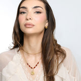 Woman wearing an 18k gold plated layered necklace with a ruffled flower pendant and dark reddish-brown tube beads, paired with hoop earrings