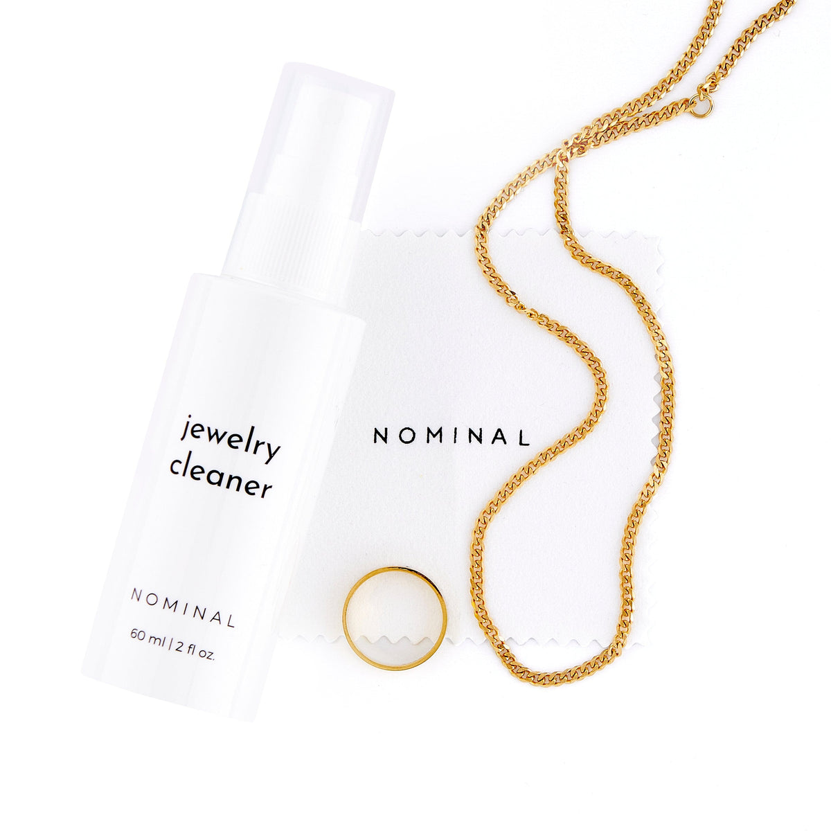 Flat lay of the Nominal Jewelry Cleaner kit, including a white spray bottle labeled “jewelry cleaner” (60 ml / 2 fl oz), a white microfiber cloth with the word “NOMINAL” printed in black, a gold chain, and a gold ring.