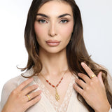 Woman wearing an 18k gold plated necklace with a detailed flower stem pendant and reddish-brown tube beads, paired with gold rings