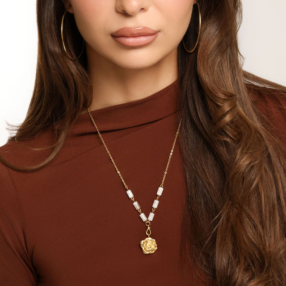 Woman wearing an 18k gold plated necklace with a detailed rose flower pendant and milky white tube beads, paired with gold hoop earrings