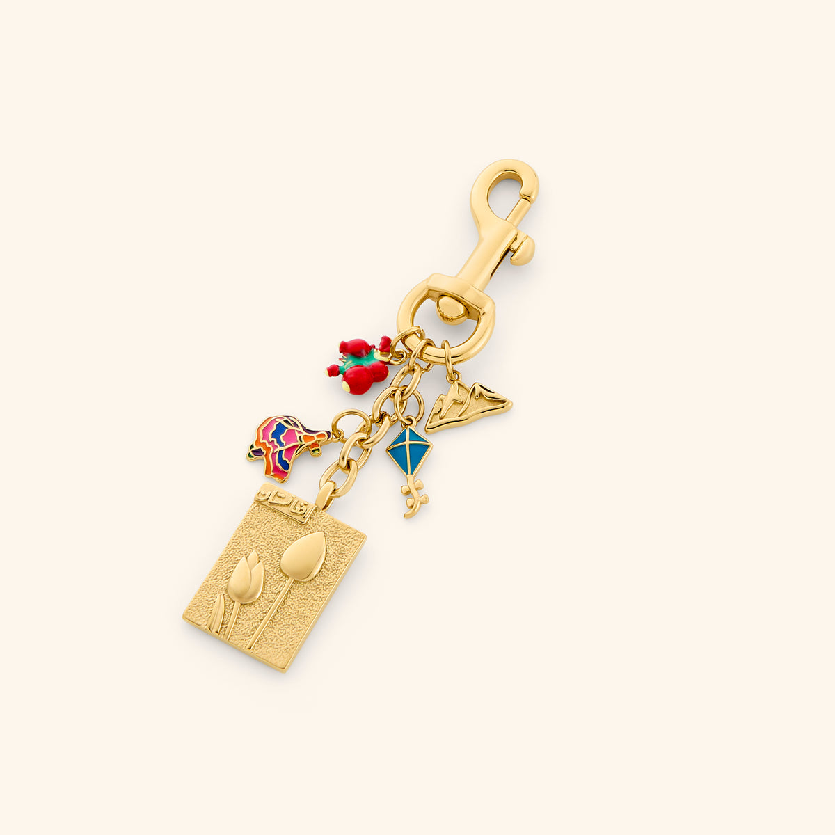 The Afghanistan Stories Bag Charm by Nominal, crafted with an 18k gold-plated finish featuring heritage charms including an embossed tulip postage stamp, blue kite, mountain silhouette, and colorful enameled motif, displayed on an off-white background.