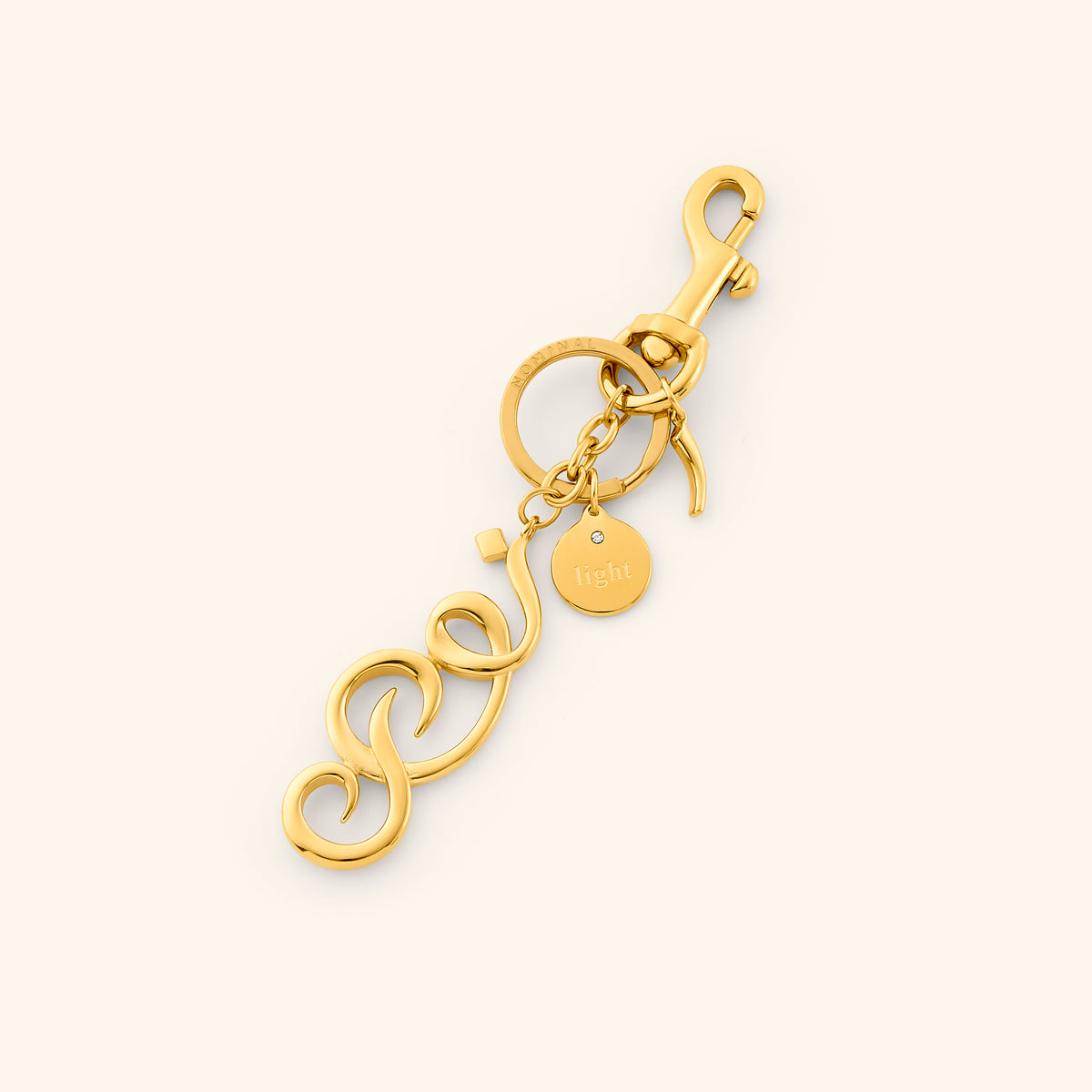 The Noor | Light Bag Charm by Nominal, crafted with an 18k gold-plated finish featuring an Arabic calligraphy charm spelling "Noor" with a swivel clasp and engraved coin tag, displayed on an off-white background