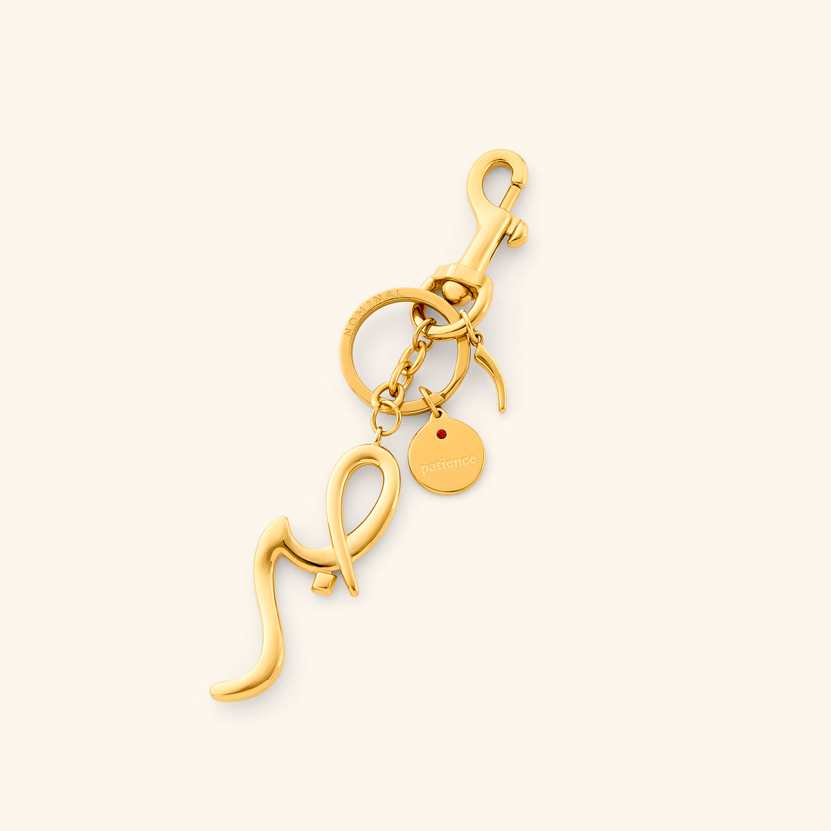 The Sabr | Patience Bag Charm by Nominal, crafted with an 18k gold-plated finish featuring an Arabic calligraphy charm spelling "Sabr" with a swivel clasp and engraved coin tag, displayed on an off-white background.