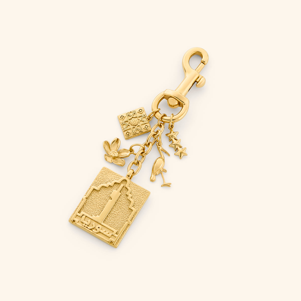 The Syria Stories Bag Charm by Nominal, crafted with an 18k gold-plated finish featuring cultural charms including an embossed Syrian architectural postage stamp, jasmine flower, and heritage pendants, displayed on an off-white background.