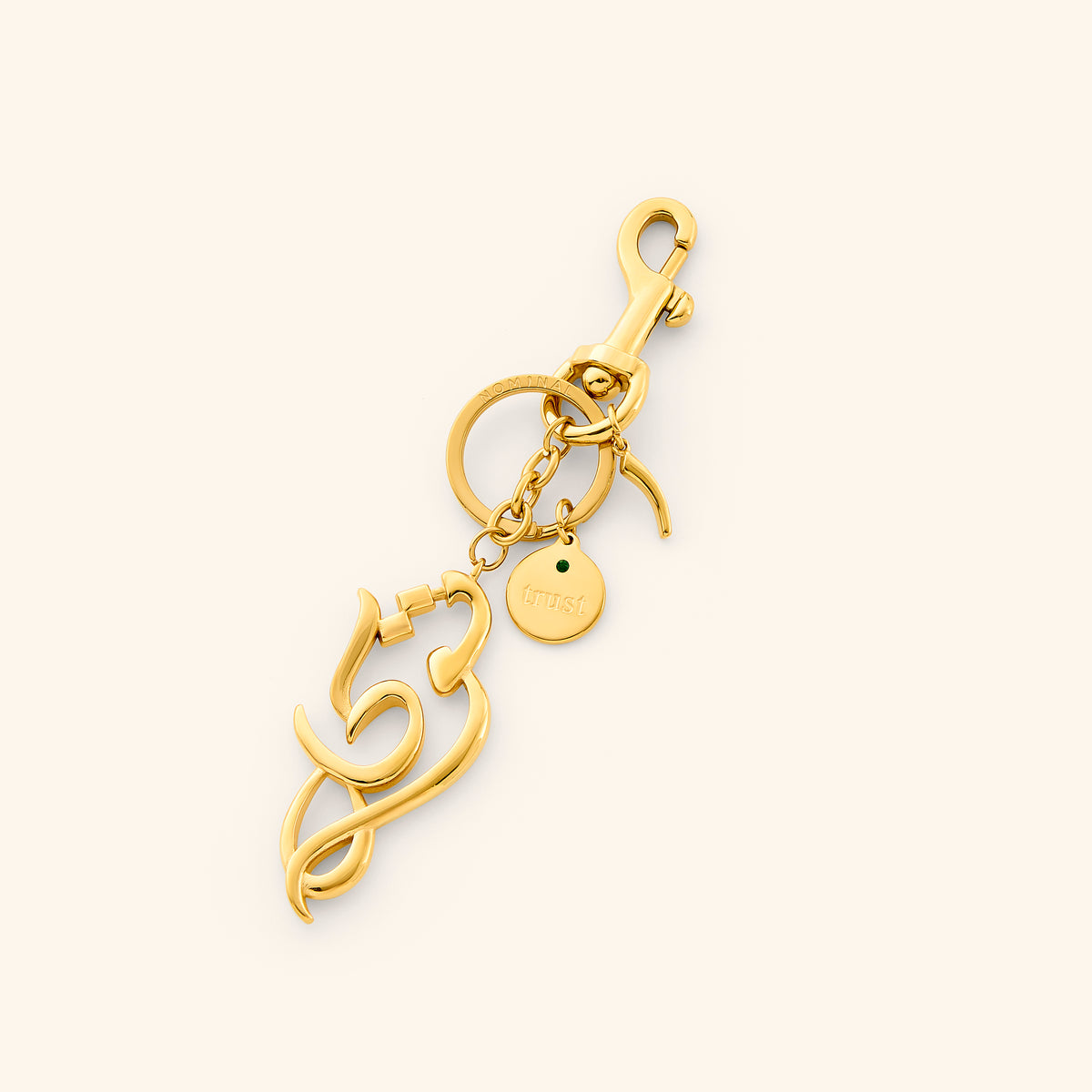 The Tawakkul | Trust Bag Charm by Nominal, crafted with an 18k gold-plated finish featuring an Arabic calligraphy charm spelling "Tawakkul" with a swivel clasp and engraved coin tag, displayed on an off-white background.