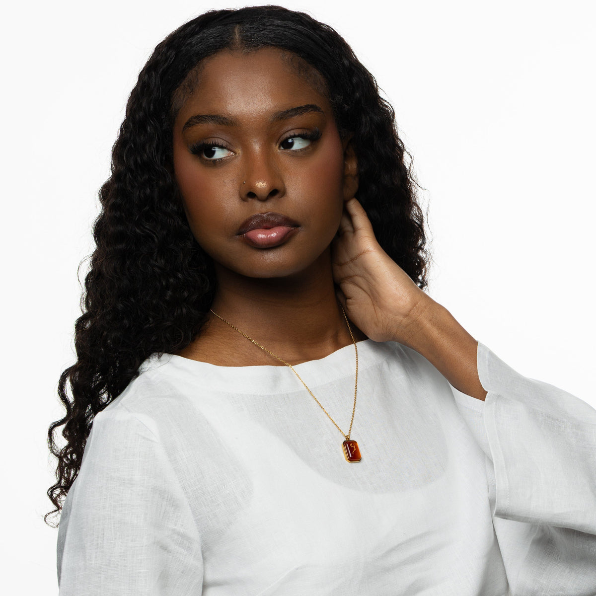 Woman wearing a gold chain necklace with a rectangular red agate stone pendant engraved with "Love" in Arabic calligraphy, styled with a white blouse, looking to the side with hand resting near neck.