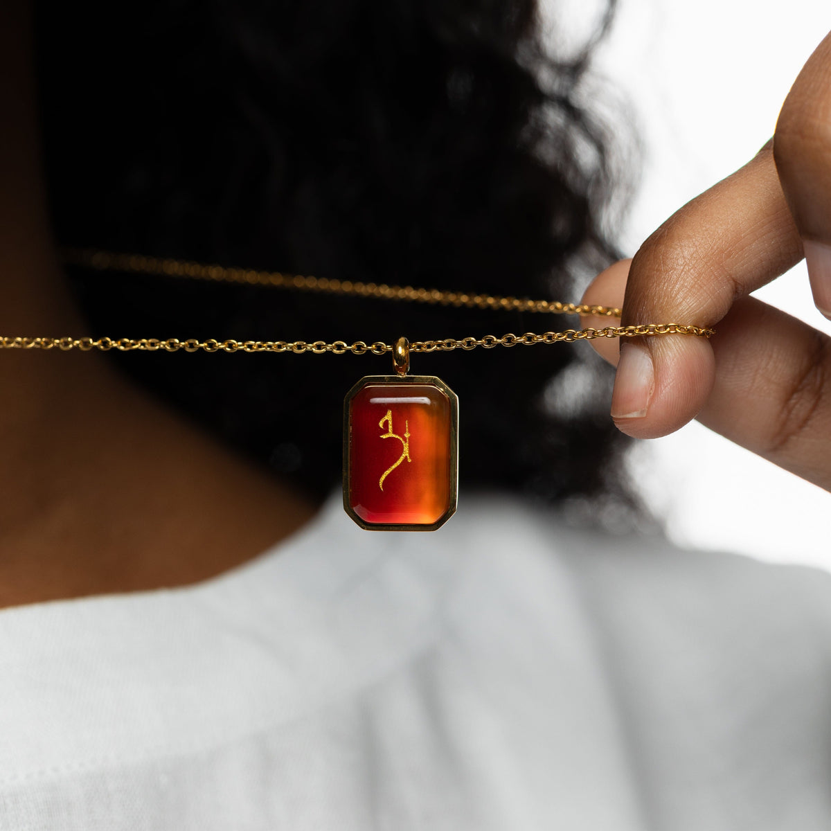 Woman holding a gold chain necklace with a rectangular red agate pendant engraved with "Love" in Arabic calligraphy, styled against a white blouse.