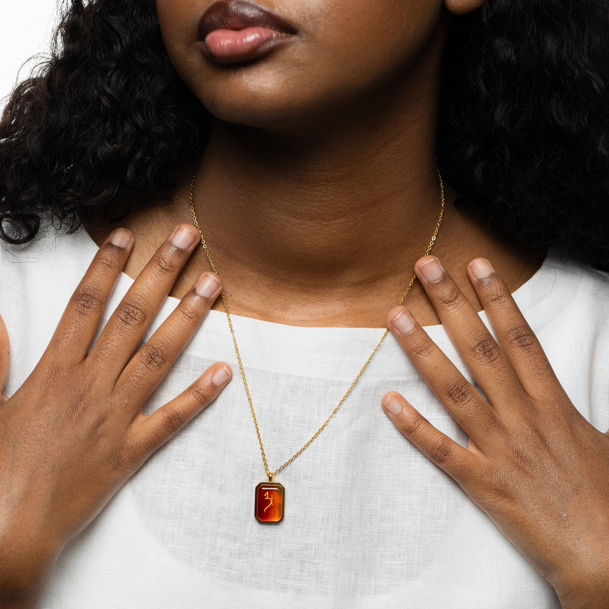 Close-up of a woman wearing a gold chain necklace with a rectangular red agate pendant engraved with "Love" in Arabic calligraphy, styled with a white blouse, both hands placed near the necklace.
