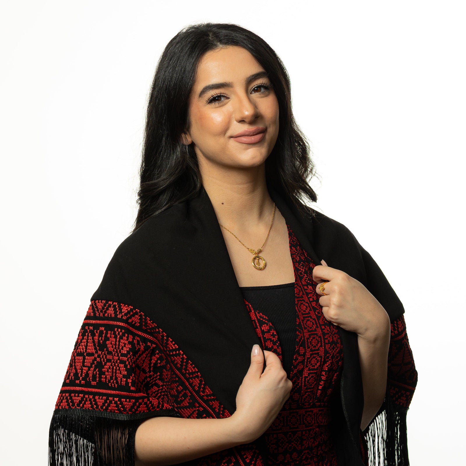 Model smiles at the camera while holding the fabric of their clothing. The model is wearing gold Love of the Homeland Lebanon Cedar Necklace around their neck and gold ring on their left middle finger.