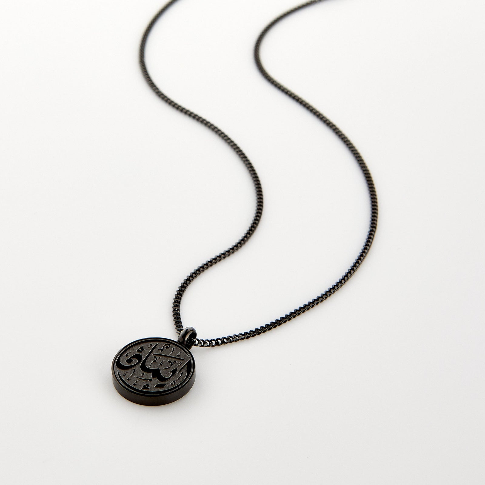 A black Faith Token necklace laid flat on a white background, showing the engraved Arabic script “Iman”.