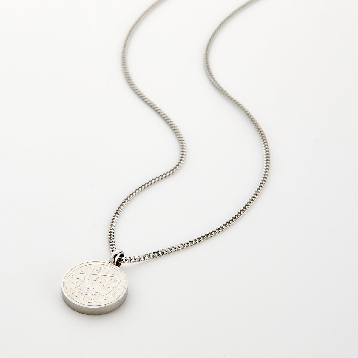 A silver Faith Token necklace with the pendant resting flat on a light background, showing the engraved Arabic script “Iman”.