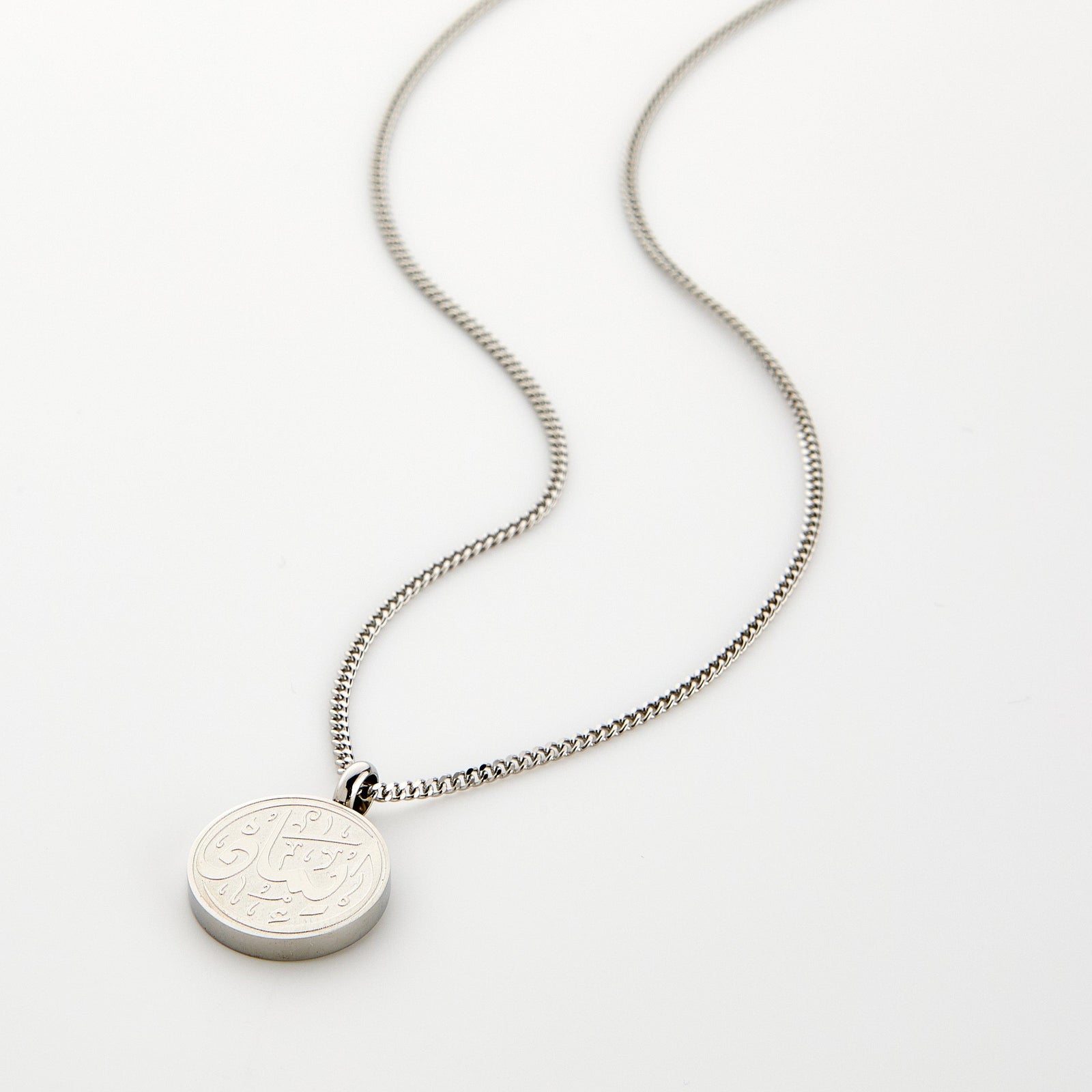 A silver Faith Token necklace with the pendant resting flat on a light background, showing the engraved Arabic script “Iman”.