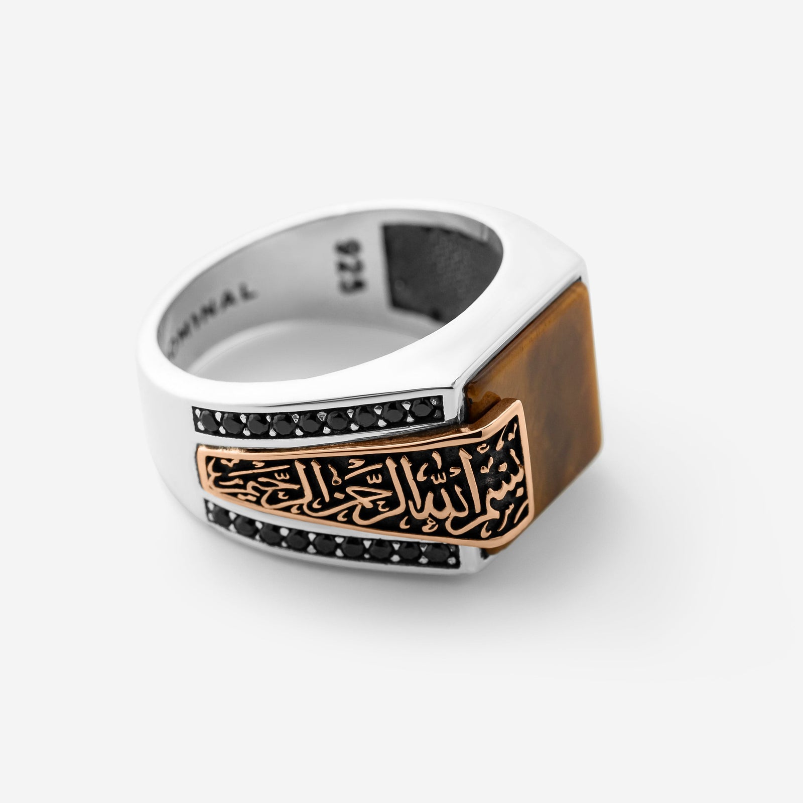 A photo of a sterling silver ring that features a striking square Tiger's Eye stone anchors this statement piece, with “بِسْمِ اللهِ الرَّحْمٰنِ الرَّحِيْم” (Bismillāhir-Raḥmānir-Raḥīm) engraved in an encased calligraphy that spills from the band onto the stone itself. On the opposite side, “الحمدُ لله” (Al-ḥamdu lillāh) is carved into the band.