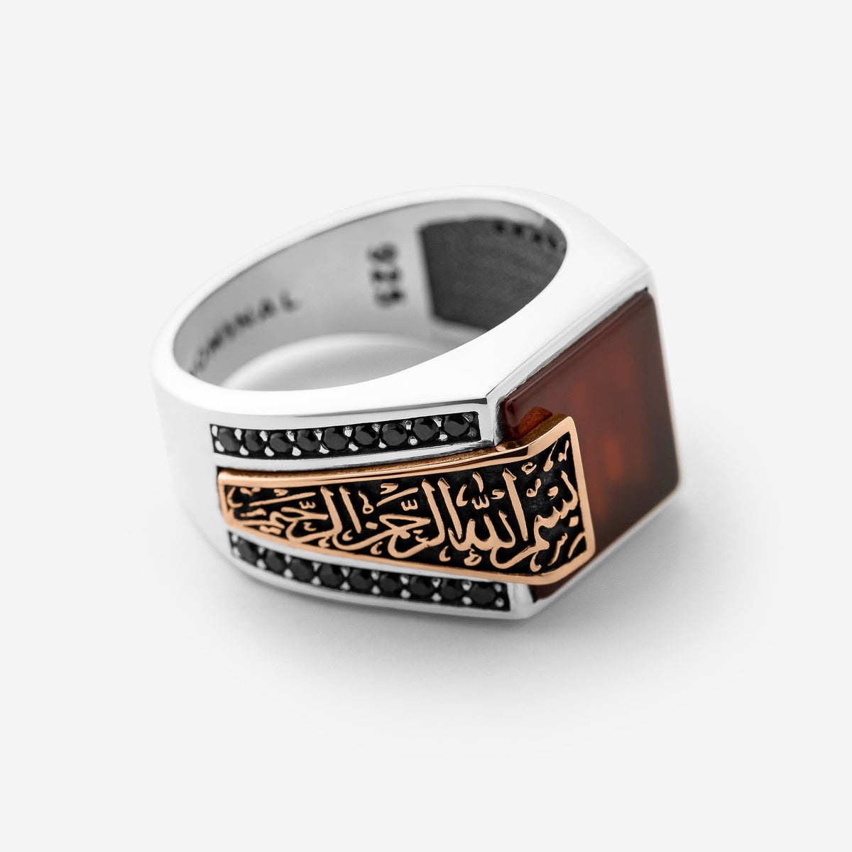 Photo of a ring that features a striking deep red agate stone stone anchors this statement piece, with “بِسْمِ اللهِ الرَّحْمٰنِ الرَّحِيْم” (Bismillāhir-Raḥmānir-Raḥīm) engraved in an encased calligraphy that spills from the band onto the stone itself.