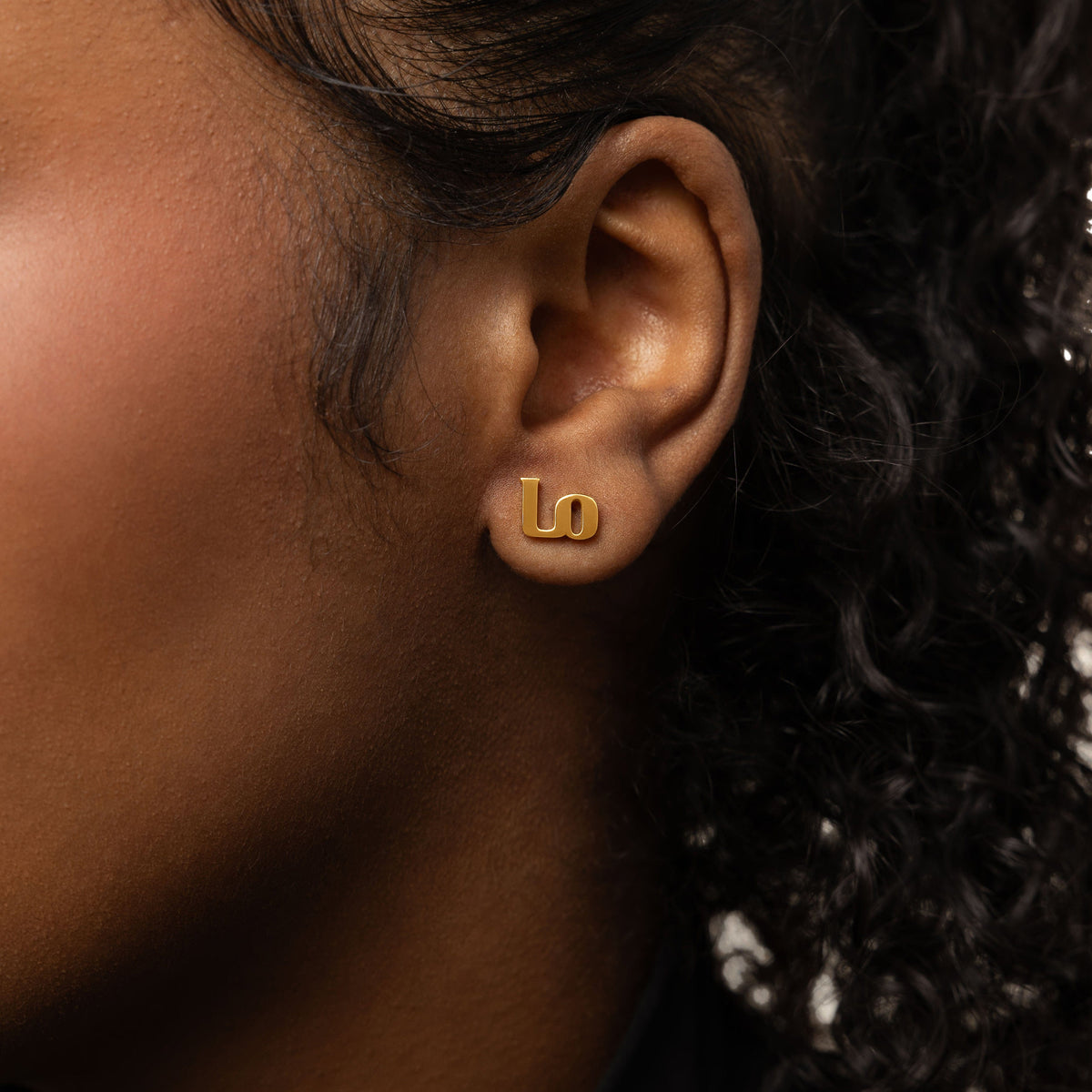 A female model with medium skin tone wearing the 18K gold-plated "Mama" stud earrings. The earrings sit delicately on her earlobes, complementing her minimalist outfit and highlighting the meaningful Arabic inscription.