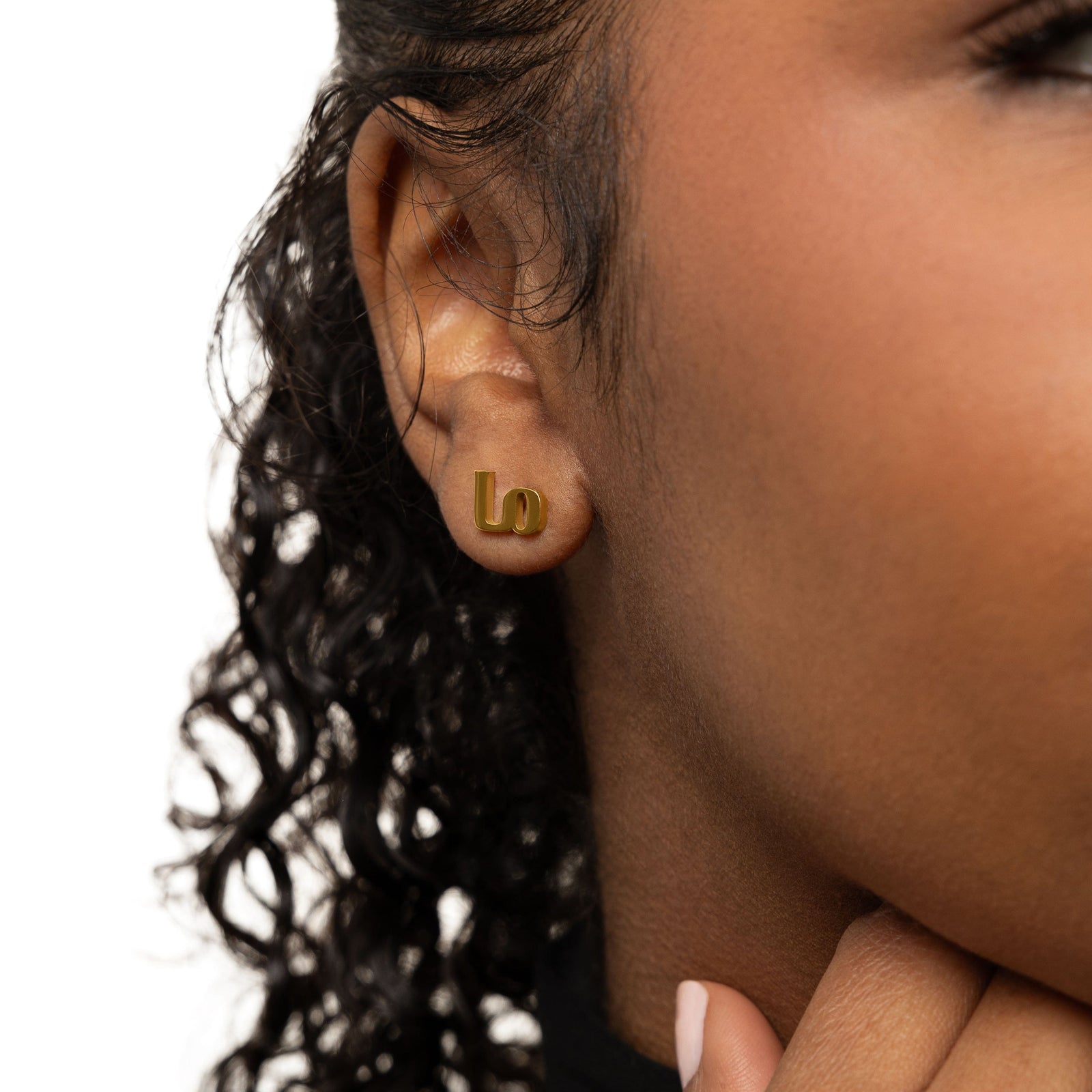 A female model with medium skin tone wearing the 18K gold-plated "Mama" stud earrings. The earrings sit delicately on her earlobes, complementing her minimalist outfit and highlighting the meaningful Arabic inscription.