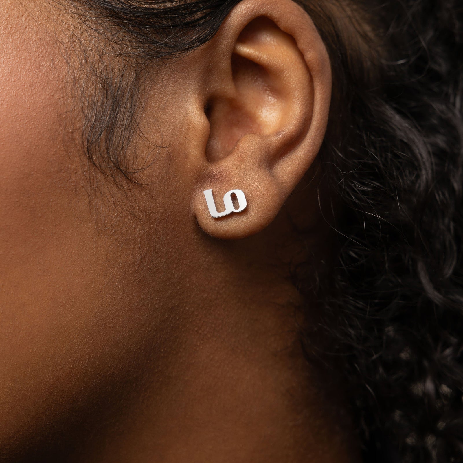 A female model with medium skin tone showcasing the silver "Mama" stud earrings. The earrings rest elegantly on her earlobes, enhancing her simple attire and drawing attention to the detailed Arabic lettering.