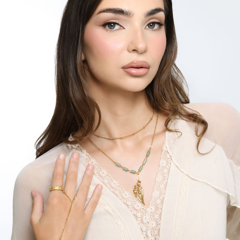 Woman wearing an 18k gold plated layered necklace with a lily of the valley pendant and pale green tube beads, paired with a gold ring and hand chain