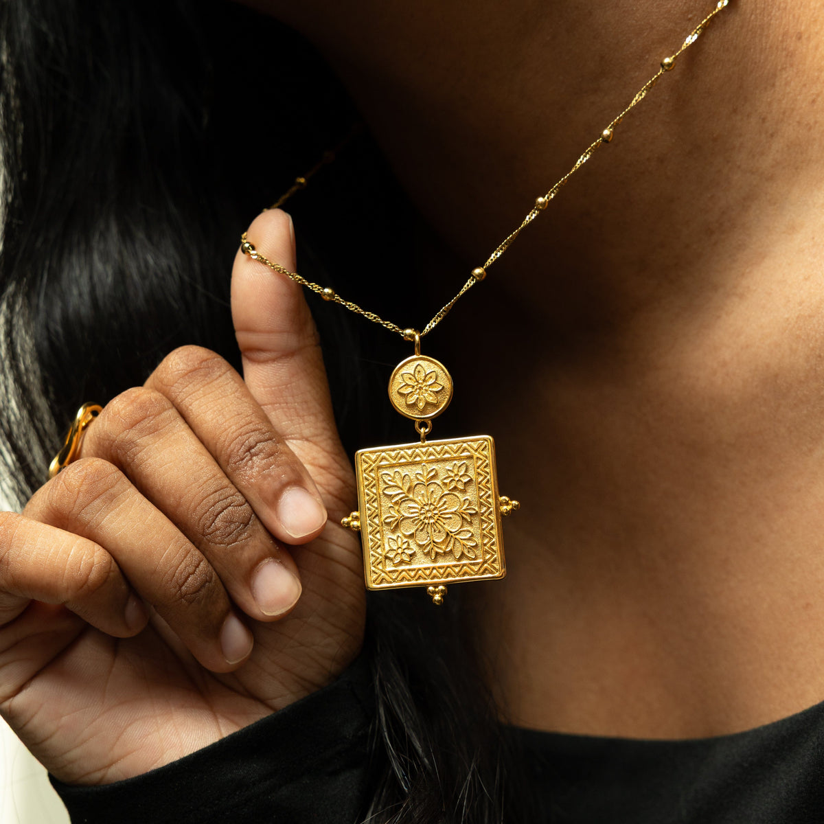 Woman holding up the Mehndi Floral Necklace, showing the gold floral pendant close to her neck.