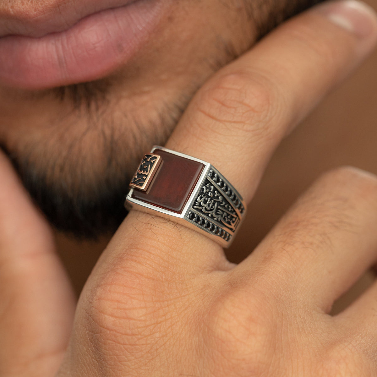 Man wearing a silver ring with a red agate stone and an Arabic calligraphy plate reading ‘Bismillah, Alhamdulillah,’ styled with black side stones.