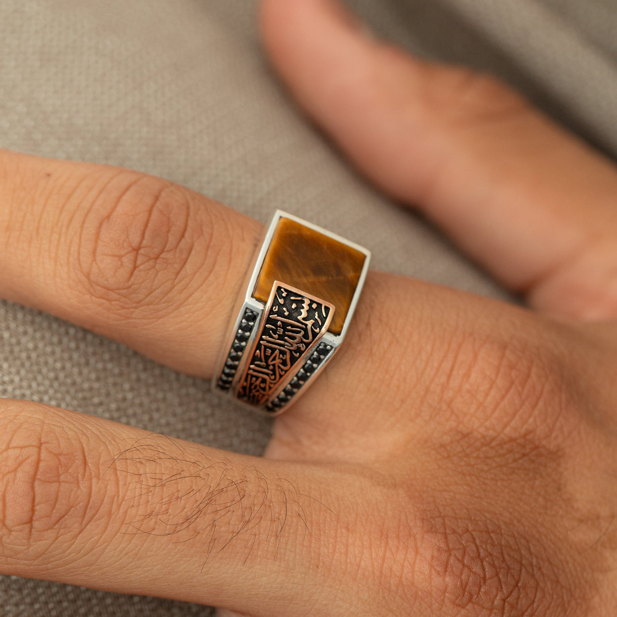 A silver ring with a tiger's eye stone and a an Arabic calligraphy plate reading ‘Bismillah, Alhamdulillah,’ styled with black side stones.