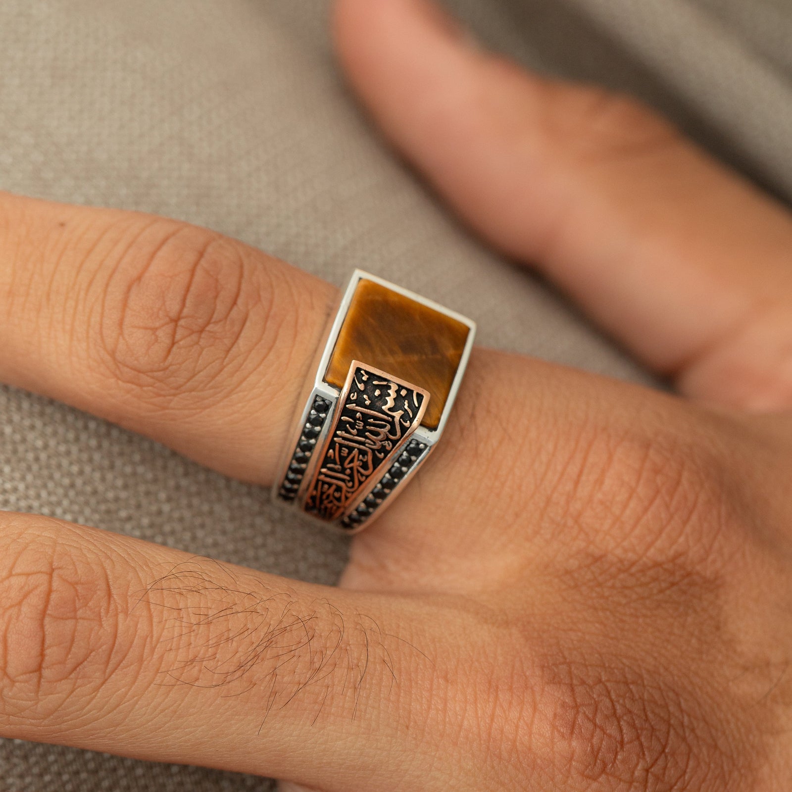 A silver ring with a tiger's eye stone and a an Arabic calligraphy plate reading ‘Bismillah, Alhamdulillah,’ styled with black side stones.