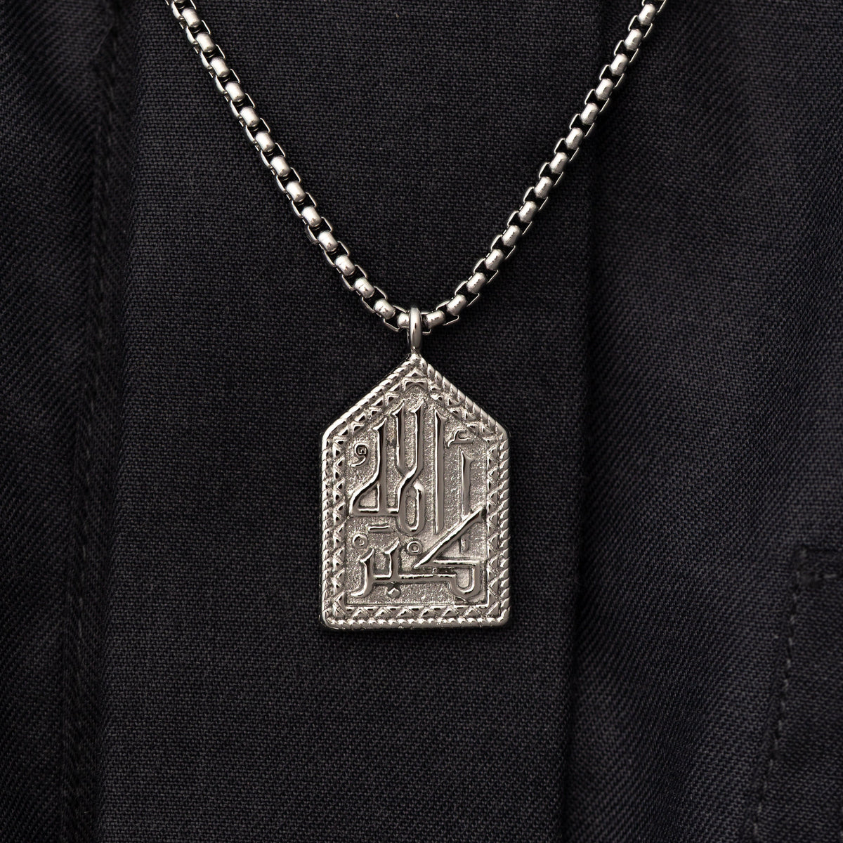 Close up of the silver "Allahu Akbar" Calligraphy Necklace on a black background.