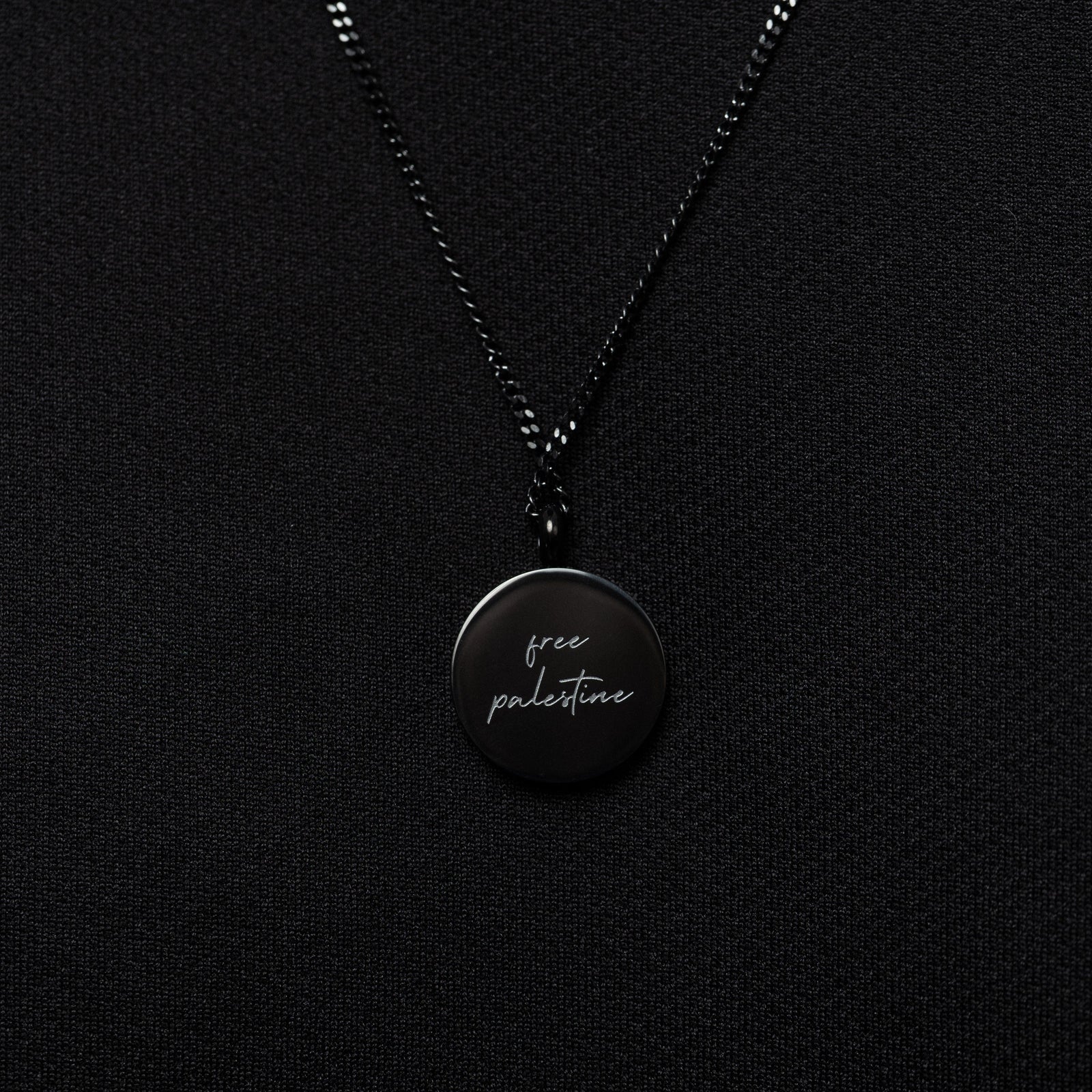 Black Al-Aqsa ‘Free Palestine’ Necklace on a black fabric, showing the back of the pendant with english text engraving that reads "Free Palestine"