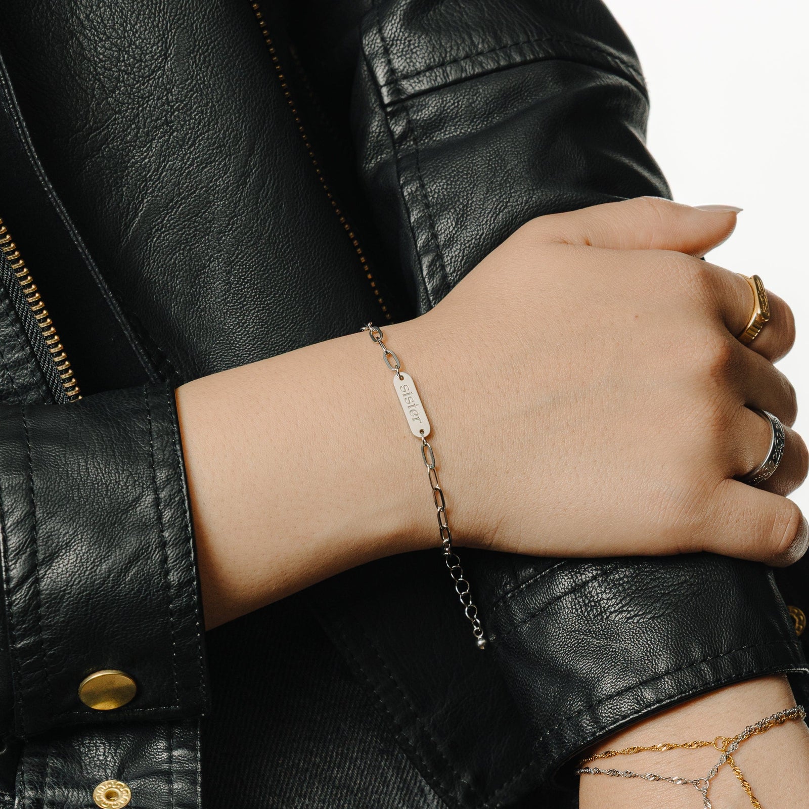 Wrist adorned with silver version of the bracelet featuring "sister" engraved in English, styled with layered rings and a black leather jacket.
