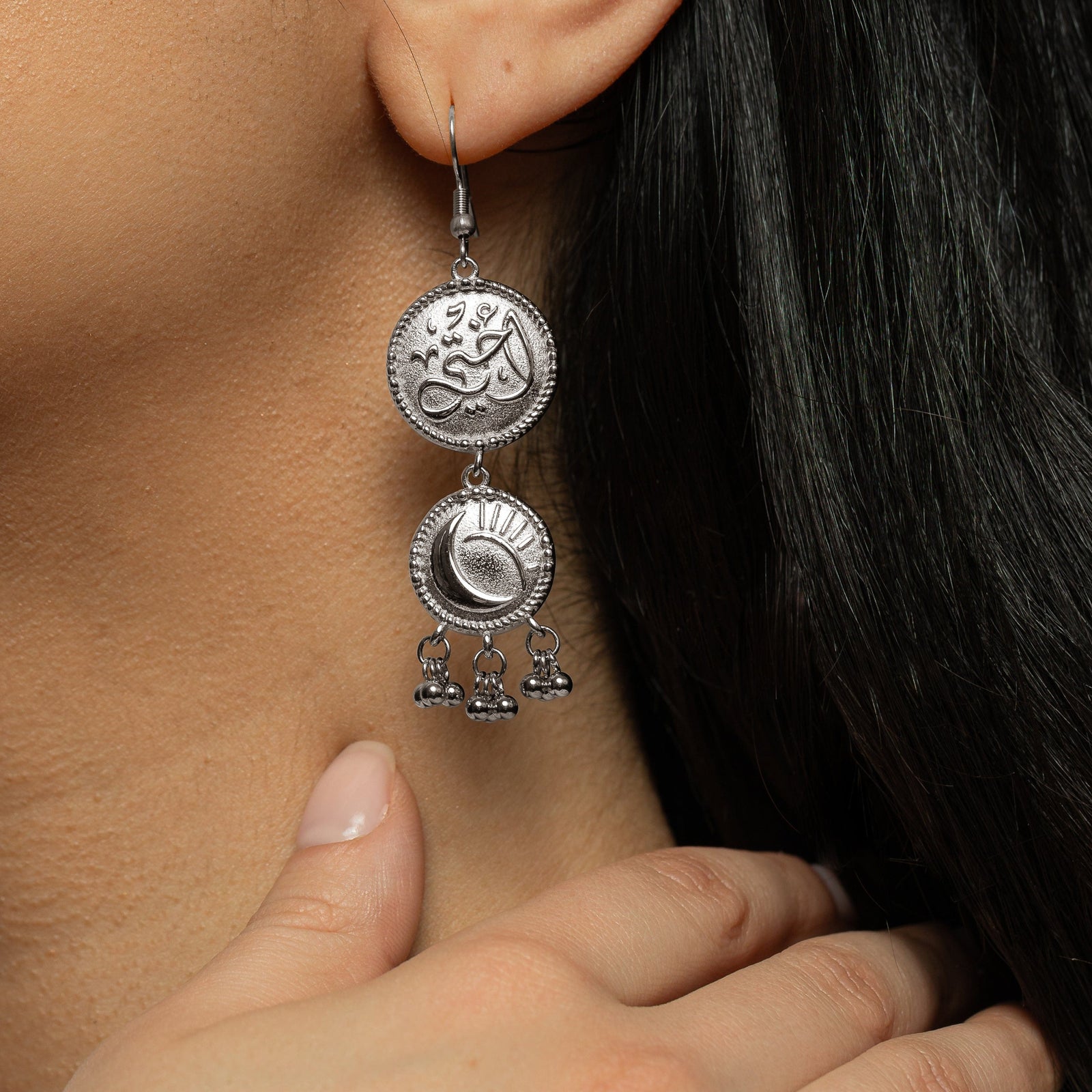 Close up photo of the silver My Sister Sun & Moon Coin Earring as it dangles on the model's left ear. Model's hand placed on the side of their neck just above the left shoulder.