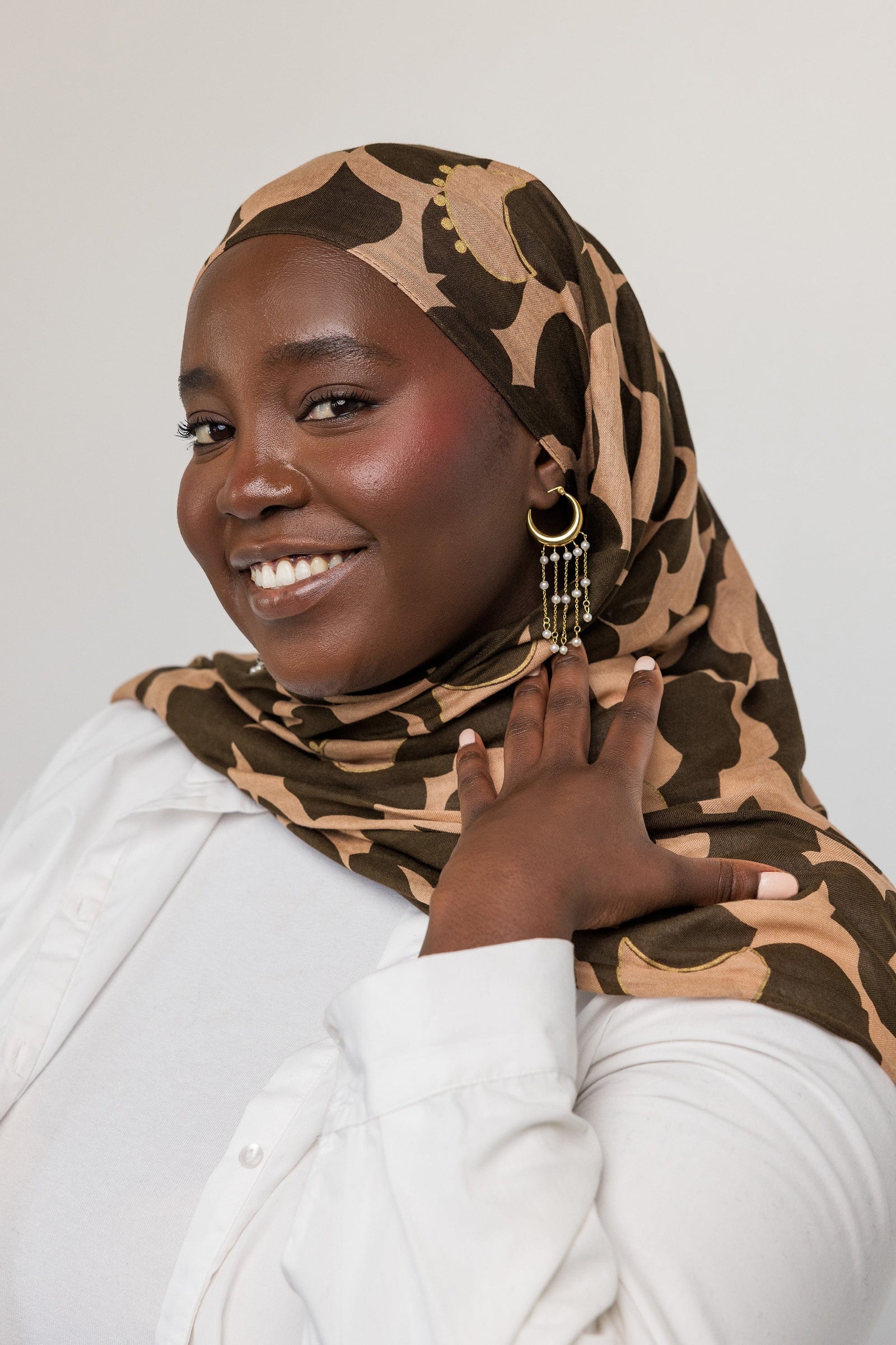 Photo of the model smiling to the camera. The model is touching their hijab to show the gold Raining Pearl Hoop earring they are wearing on their left ear.