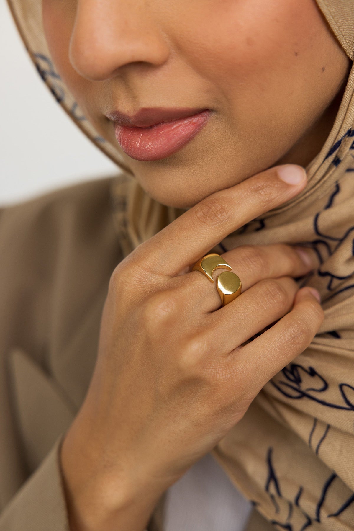 Woman wearing the Gold Phases Ring on her finger, styled with a beige patterned hijab and tan blazer, hand resting on her chin.