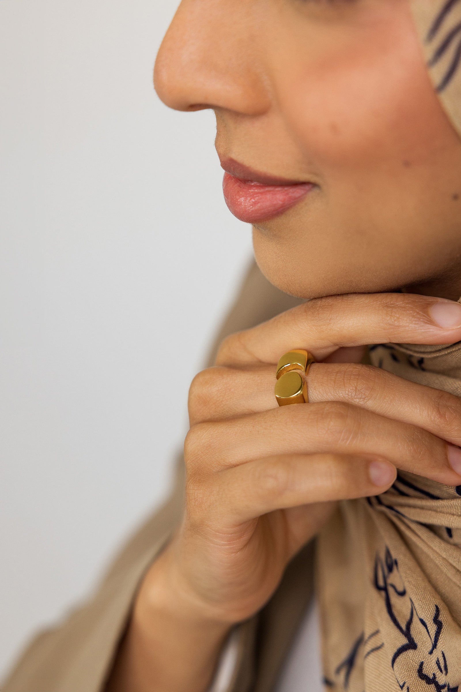Woman wearing the Gold Phases Ring on her finger, styled with a beige patterned hijab and tan blazer, hand resting against her chin.
