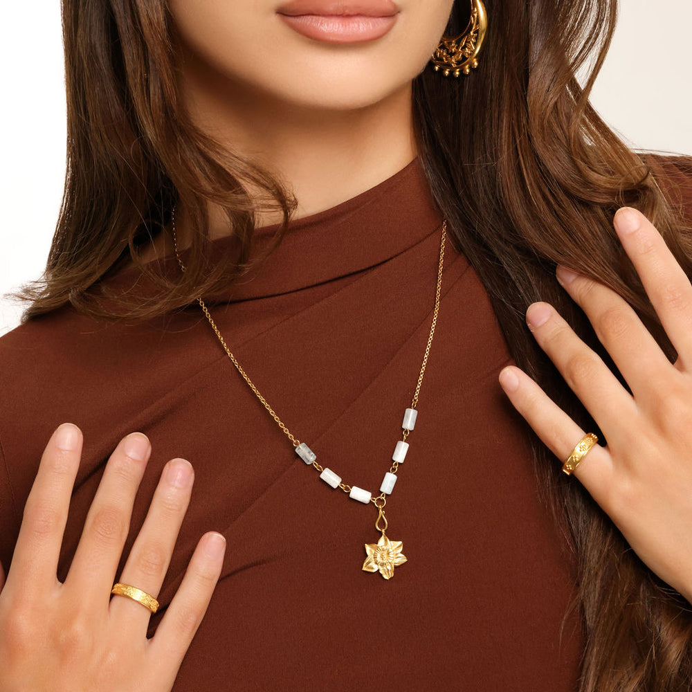 Woman wearing an 18k gold plated necklace with a detailed flower pendant and pale blue tube beads, paired with gold hoop earrings and rings