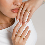 Model wearing 18k gold-plated jewelry featuring two ring stacks: on the upper hand, the Mughal Bloom Bezel Ring on the index finger, Intertwined Bezel Ring on the middle finger, and North Star Bezel Ring on the ring finger; on the lower hand, the Olive Branch Marquise Bezel Ring on the middle finger and a Five-Stone Bezel Ring stacked above a Single Bezel Ring on the ring finger.