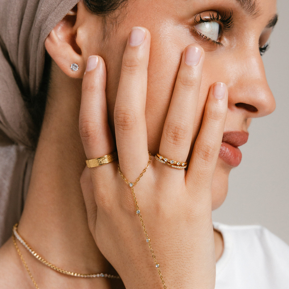 Model wearing 18k gold-plated jewelry featuring a Five-Stone Bezel Ring stacked above a Bezel Band on the index finger, a hand chain with bezel-set clear crystals anchored on the middle finger, an engraved gold band ring on the ring finger, and a Bezel Stud earring.