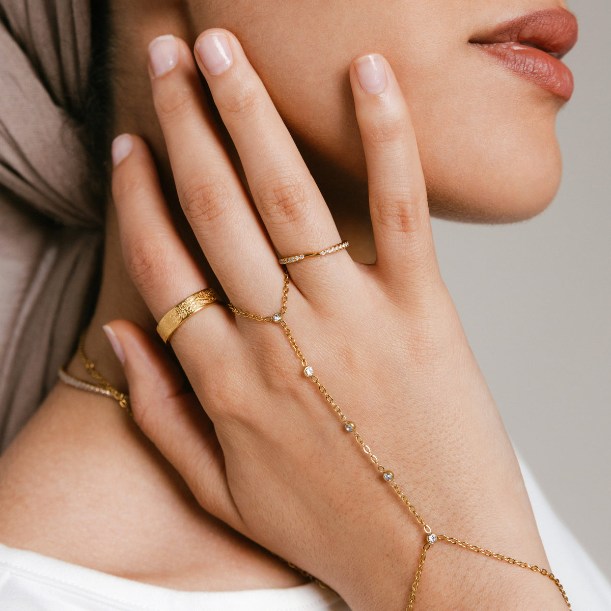 Model wearing 18k gold-plated jewelry featuring an engraved gold band ring on the index finger, a Bezel Band on the middle finger, and a hand chain with bezel-set clear crystals anchored on the middle finger.