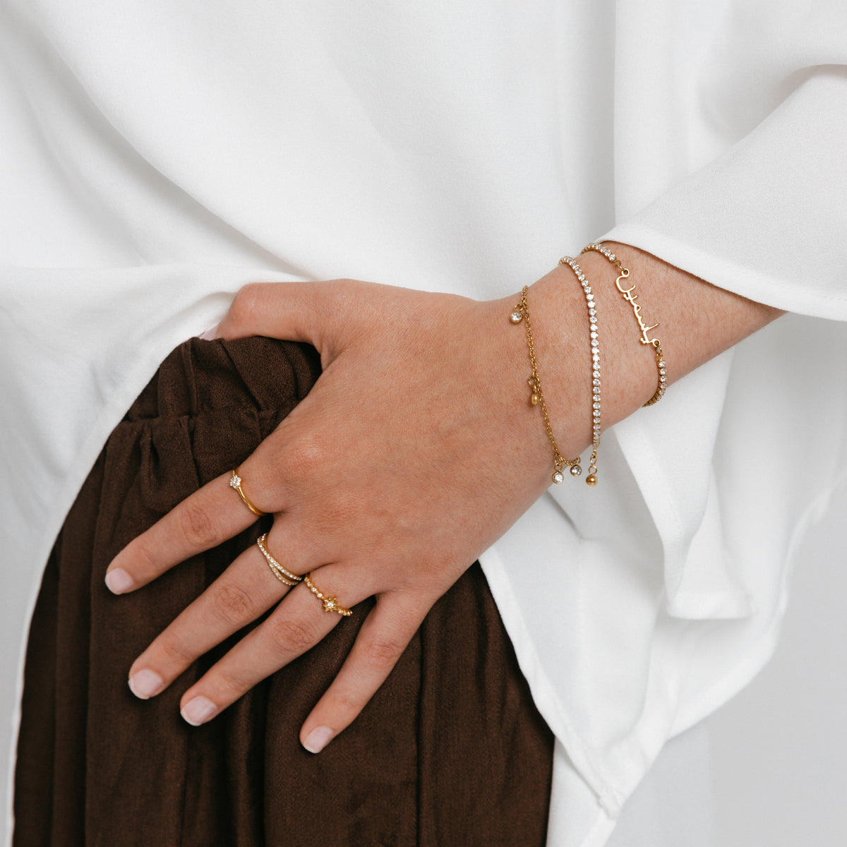 Model wearing 18k gold-plated jewelry featuring a ring stack with the Mughal Bloom Bezel Ring on the index finger, Intertwined Bezel Ring on the middle finger, and North Star Bezel Ring on the ring finger, paired with a wrist stack featuring the Tennis Name Bracelet, Tennis Bracelet, and a dangling charm bracelet.