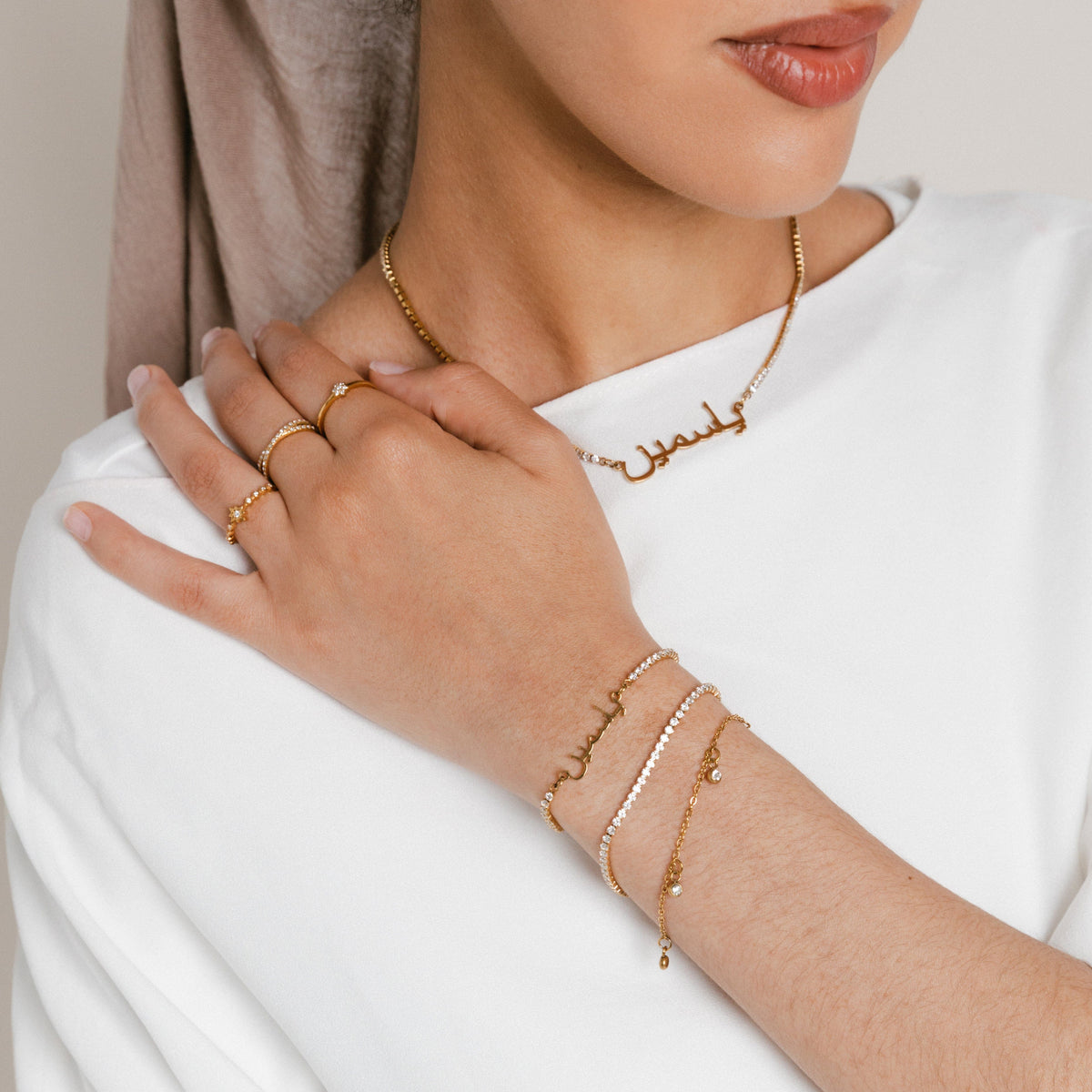Model wearing a jewelry set featuring 18k gold-plated pieces: a ring stack with the Mughal Bloom Bezel Ring on the index finger, Intertwined Bezel Ring on the middle finger, and North Star Bezel Ring on the ring finger, paired with a custom Arabic calligraphy Tennis Name Necklace and a wrist stack featuring the Tennis Name Bracelet alongside a pavé crystal tennis bracelet and a delicate charm bracelet.